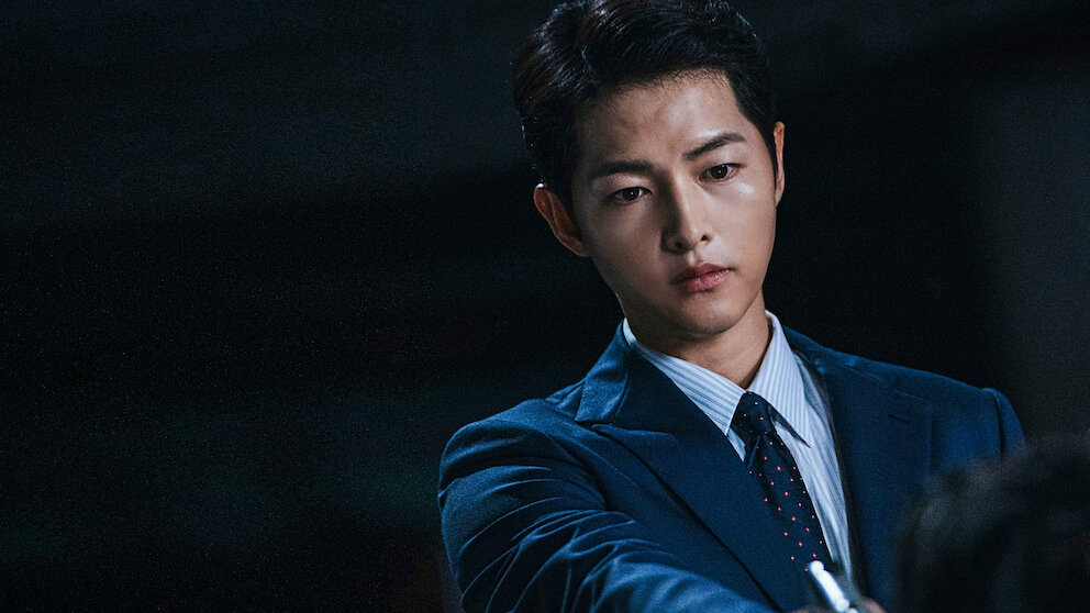Song Joong-ki em cena de Vincenzo, vestindo terno escuro em ambiente com pouca luz.